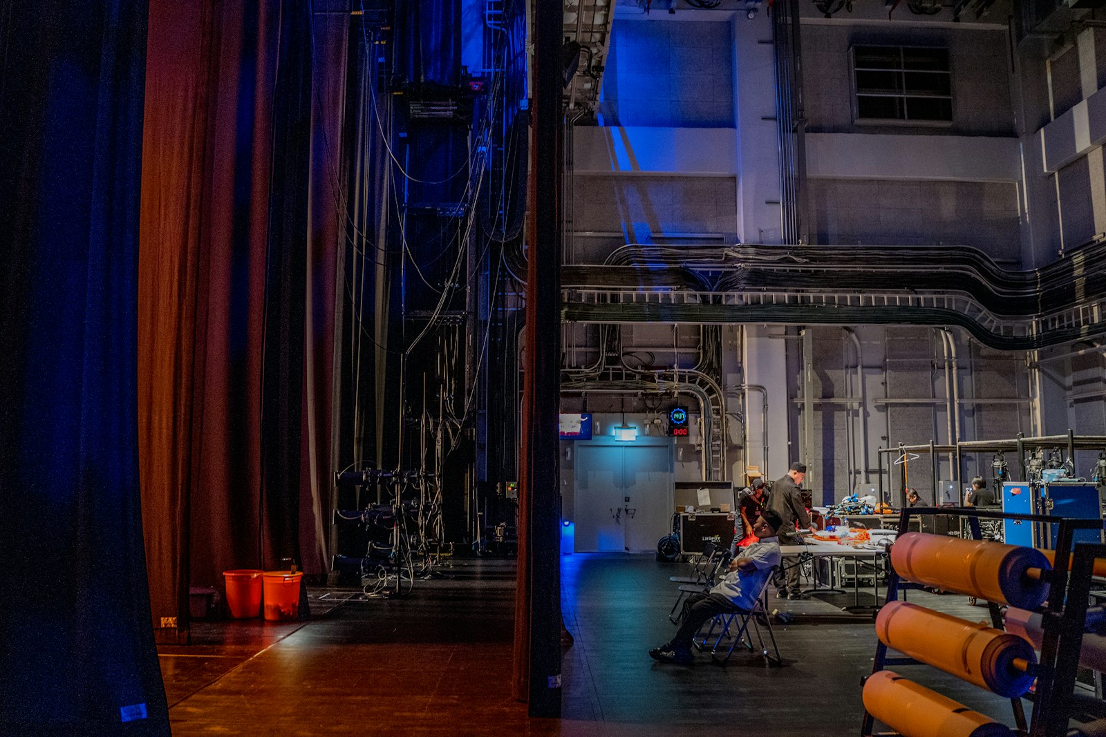 Crew working backstage behind a parted stage curtain among rigging, cables and flight cases.