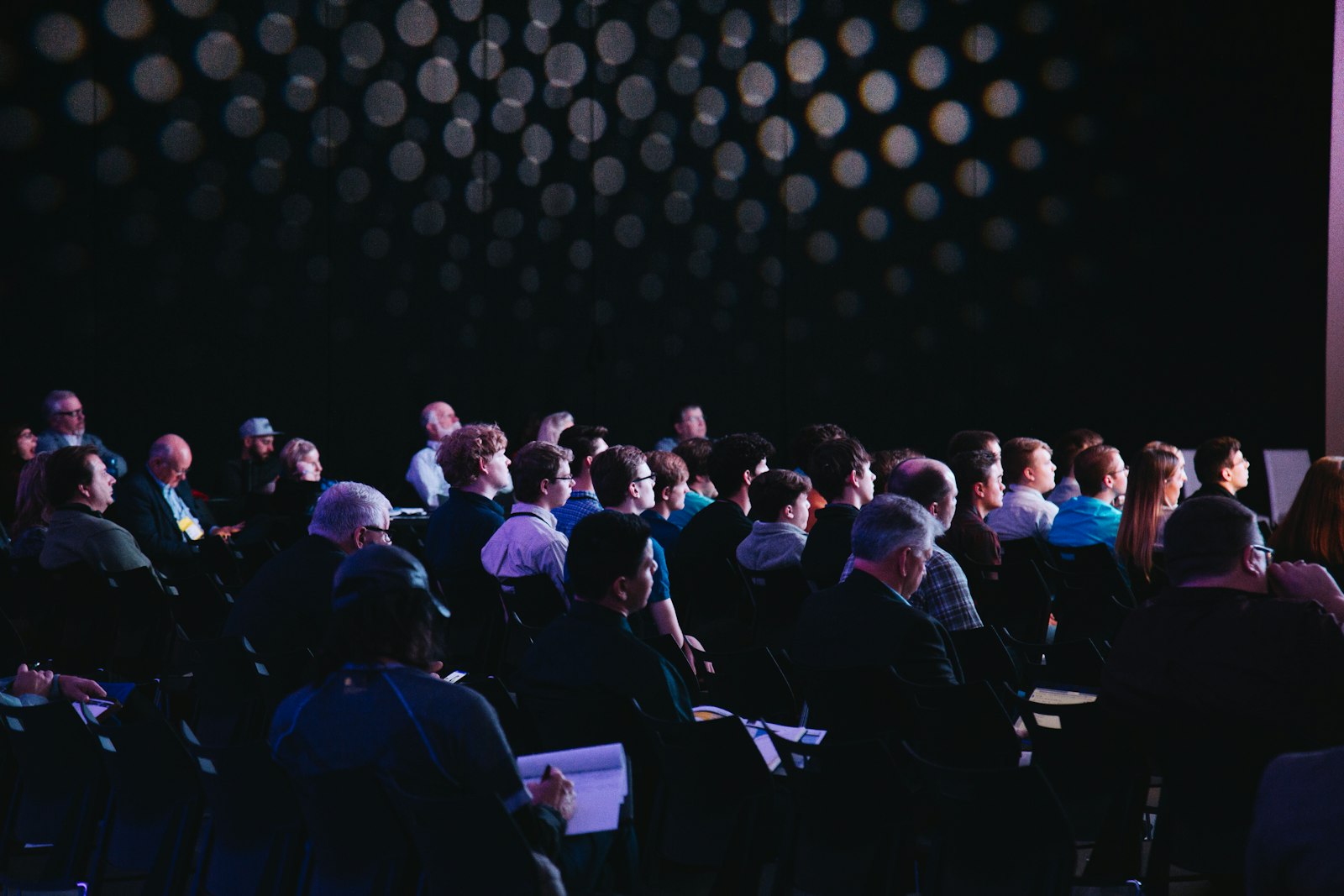 A seated conference audience watching a session in a darkened hall with a bokeh light wall behind them.