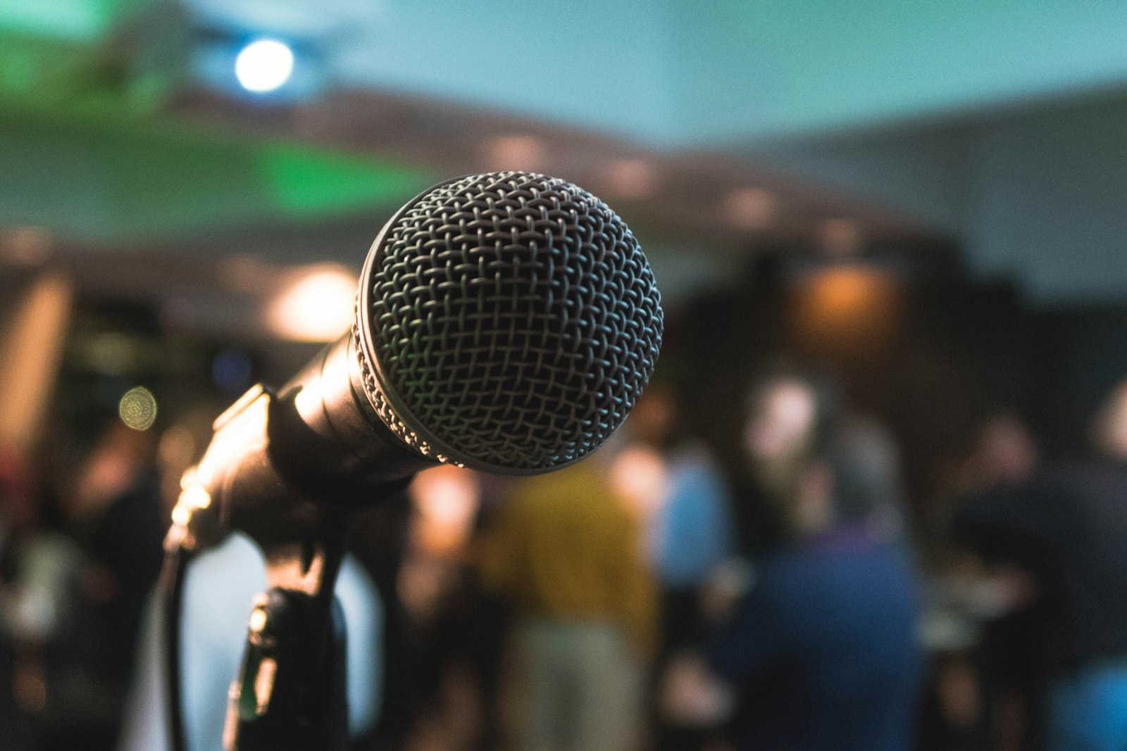 A stage microphone in sharp focus with a softly blurred audience behind it.