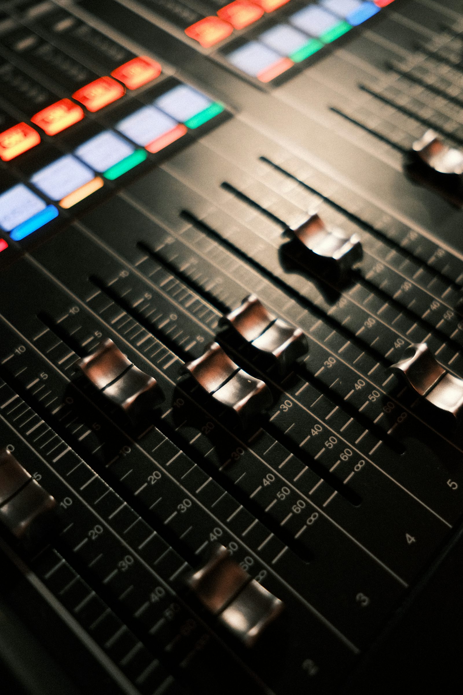A close crop of a broadcast mixing console: angled metal faders and small illuminated buttons in low light.