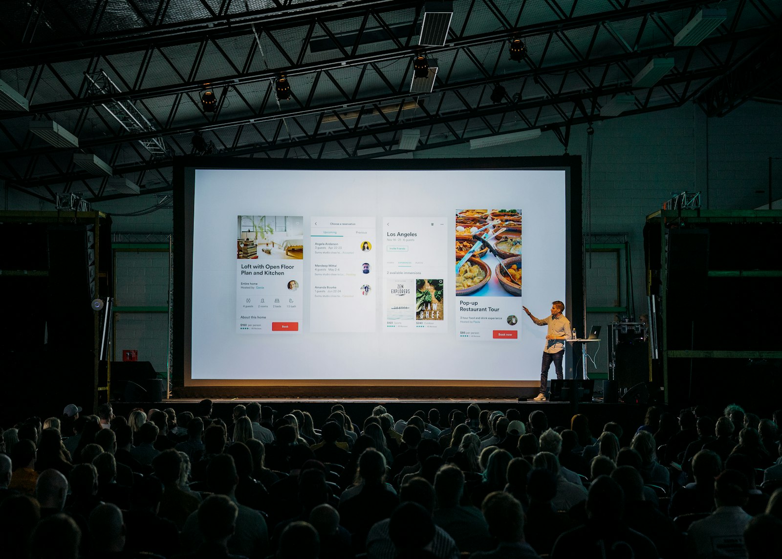 A speaker presenting product screens on a large stage screen to a seated audience.